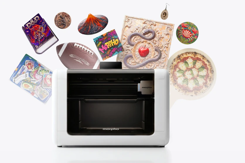 The Morpho UV printer surrounded by its sample prints: a textured D&D book cover, the 35 mm volcano, an embossed relief panel, stickers, an earring and a printed football